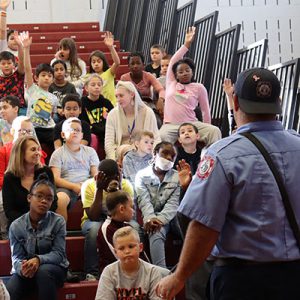 A firefighter calls on students seated in the bleachers with their hands raised to ask questions.