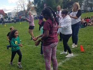 A Watervliet Elementary School student ambassador hands a student an apple at the finish line. A teaching assistant, kindergarten teacher, the principal and coach applaud two students crossing the finish line. 