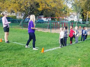 A group of kindergarten students stand at the starting line while a teacher stands nearby and the coach readies the whistle to begin the Apple Run.