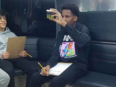 One student examines a water sample in a clear plastic cup while another student holding a clip board looks on during the Bio Bus filed trip. 