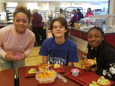 Three high school students seated in the cafeteria. All have pink face paint, and smiling at the camera. One student, wearing a pink top, is holding the face paint. 