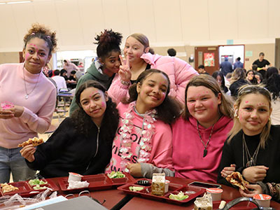 Group of high school students, most wearing pink clothing, seated at cafeteria table. Two students stand behind, one making a peace sign with hands. All smiling at the camera.