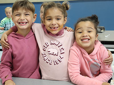 Three elementary students seated at a table, arm in arm and dressed in pink, smiling at the camera.