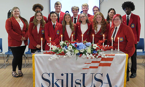 Fourteen student officers dressed in red Skills USA jackets stand behind a table draped with a Skills USA banner. The students are looking at camera and smiling.