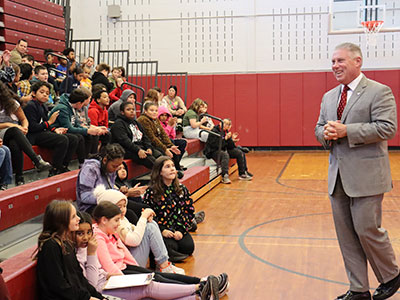 Assembly member John McDonald smiles while standing in front of fifth grade classes that are seated on the bleachers in the elementary school gym.