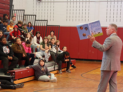 Assembly member John McDonald reads from a book as students seated in the bleachers look toward him.