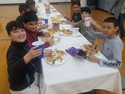 A group of students seated at a table with plates full of food, holding up paper cups and smiling at the camera.