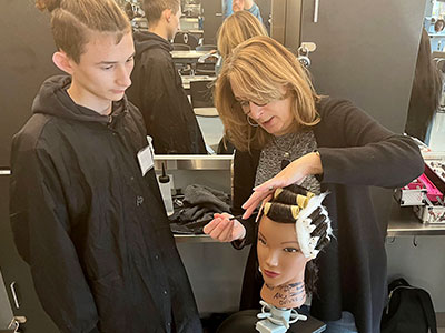 Cosmetology teacher demonstrates a technique for cutting hair using a comb and scissors while a student observes. 
