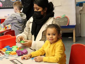 Prekindergarten student dressed in a yellow sweatshirt is seated at a desk playing with blocks and looking at the camera.