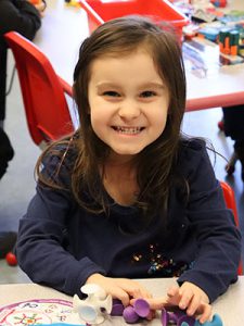 Prekindergarten student dressed in navy blue is seated at a desk and smiling at the camera.
