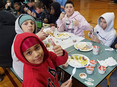 Four students seated at a table with plate of tacos in front of them. Students are smiling at the camera with thumbs up.