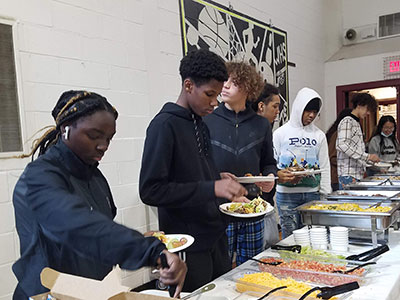Several students are lined up at serving dishes on a table loading taco meat, tortillas, vegetables, salsa cheese and guacamole onto plates.