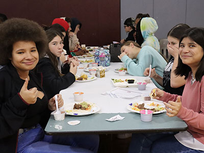 A group of students seated at a table eating tacos. Two students at the end of the table are smiling at the camera.