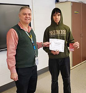 Teacher and club advisor hands award certificate to a Chess Club tournament champion who is holding a gift card prize. Both are looking at camera.