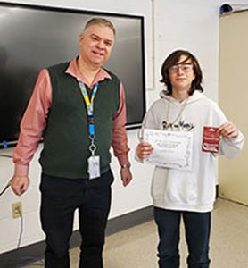 Teacher and club advisor hands award certificate to a Chess Club tournament champion who is holding a gift card prize. Both are looking at camera.