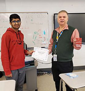 Teacher and club advisor hands award certificate to a Chess Club tournament champion who is holding a gift card prize. Both are looking at camera.