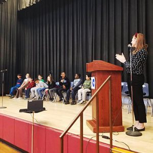 The assistant principal stands at the podium and applauds the students remaining on stage as they prepare for the next round of the spelling bee.