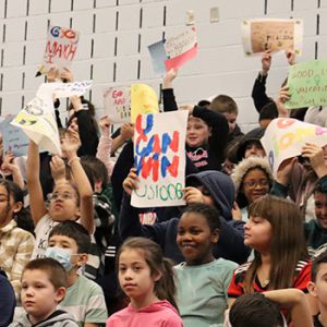 Students in the bleachers hold up signs of encouragement and cheer on their classmates during the spelling bee.