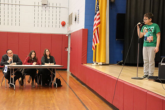 A student stands on stage at the mic spelling a word as the judges watch. 