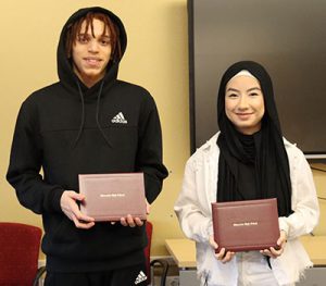 Two high school students stand side by side holding their diplomas.