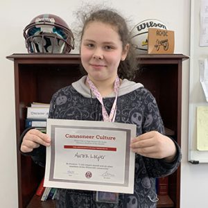 Grade 6 Student of the Month smiles at camera while holding Cannoneer Club Award Certificate