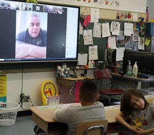 Students seated at desks listen as author on screen reads story.