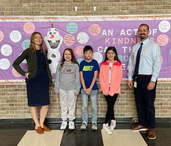 Three fifth-grade students whose essay were selected for prizes are pictured standing between assistant principal and the principal in front of a bulletin board in the elementary school lobby.