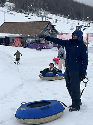 A student holding the strap attached to a snow tube points up the hill. In the background, one student seated on a snow tube is pulled through the snow by another student.