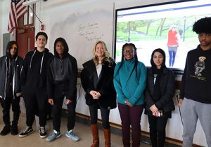 Students stand on either side of the guest speaker in front of the classroom. All are smiling at the camera.