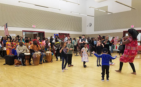 A parent leads a group of students in a traditional West African dance as three musicians play the drums in the background.
