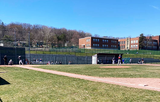 Wide shot of players standing on the baseball field with the high school in the background. 