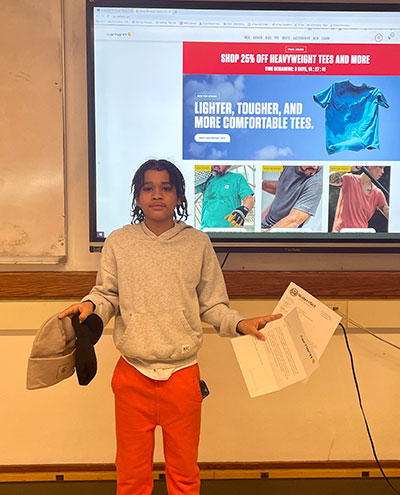 Student holds letters in one hand and Carhartt hats in the other. Behind the student is a whiteboard with the Carhartt website projected on it.