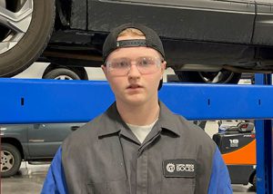 Student wearing baseball cap backwards, stands in front of a vehicle that is up on a lift.