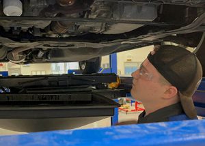 Student wearing baseball cap backwards, stands beneath a vehicle that is up on a lift and prepares to work on the engine