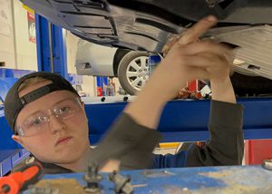 Student wearing baseball cap backwards, stands beneath a vehicle that is up on a lift and prepares to work on the engine 