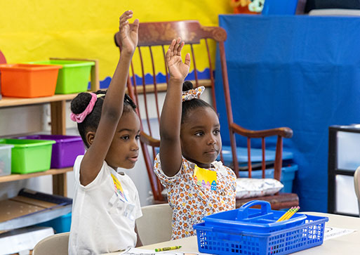 Two elementary students sit at their desks looking ahead with hands raised