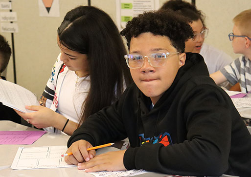 Middle school student seated at desk holding pencil smiles at the camera