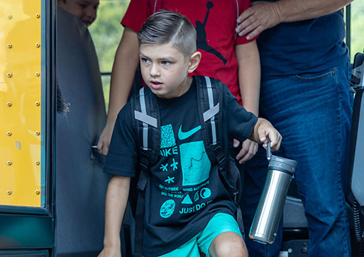 Elementary student dressed navy blue t-shirt and aqua shorts holding water bottle getting off the school bus
