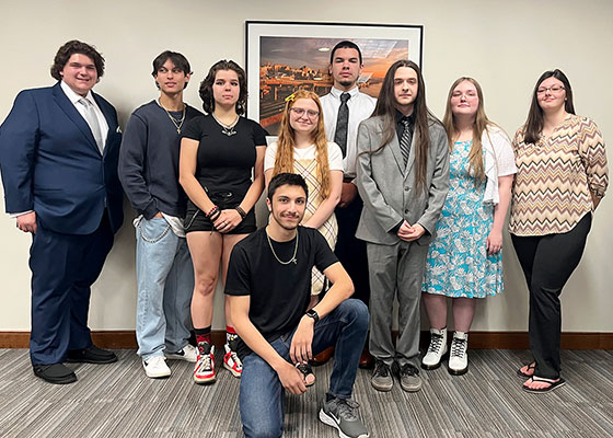 Nine students enrolled in P-TECH program stand together smiling at camera for a group photograph.  