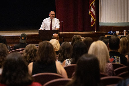 Superintendent stands at podium and speaks to teachers, staff seated in school auditorium.