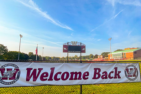 Welcome Back banner on a fence in front if the athletic field with blue sky, scoreboard and school building in the background.