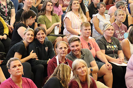 Group of teachers and staff seated on bleachers in school gymnasium. 