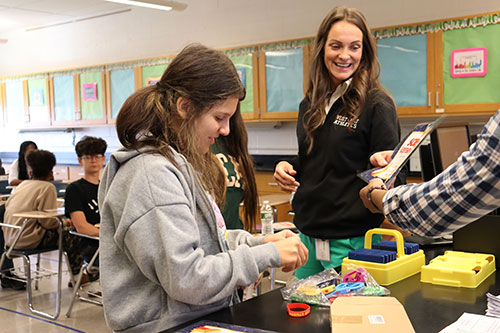 Assistant superintendent presents a certificate to a student. Another students selects a wristband at the teachers desk.