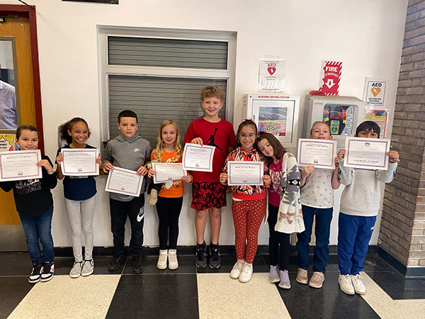 Nine elementary students stand in the hallway side-by-side holding award certificates and smiling at the camera.