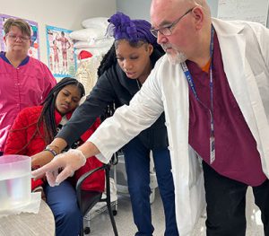 Student stands to the right of the teacher as they both point to a measuring cup that is filled with liquid. 