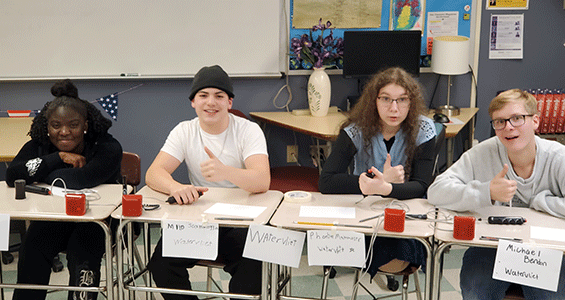 Four members of the Watervliet Master Minds team are seated at desks during the match. All are looking at the camera and smiling.
