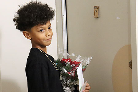 A student stands ready to knock on the door of an apartment holding a red poinsettia plant and greeting cards 