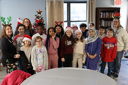The group of students and adults smile for the camera as they all stand together in front of the holiday tree in the community room of the apartment building