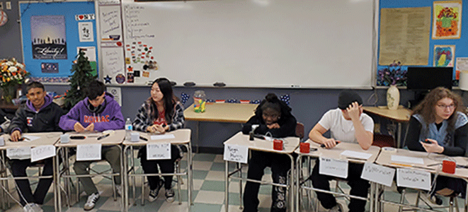 Three members from the Watervliet Master Minds team are seated at desks adjacent to the competitor's team as the match gets underway 