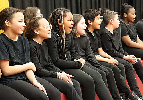 A group of students all dressed in dark clothing sits at the edge of the stage while singing 
